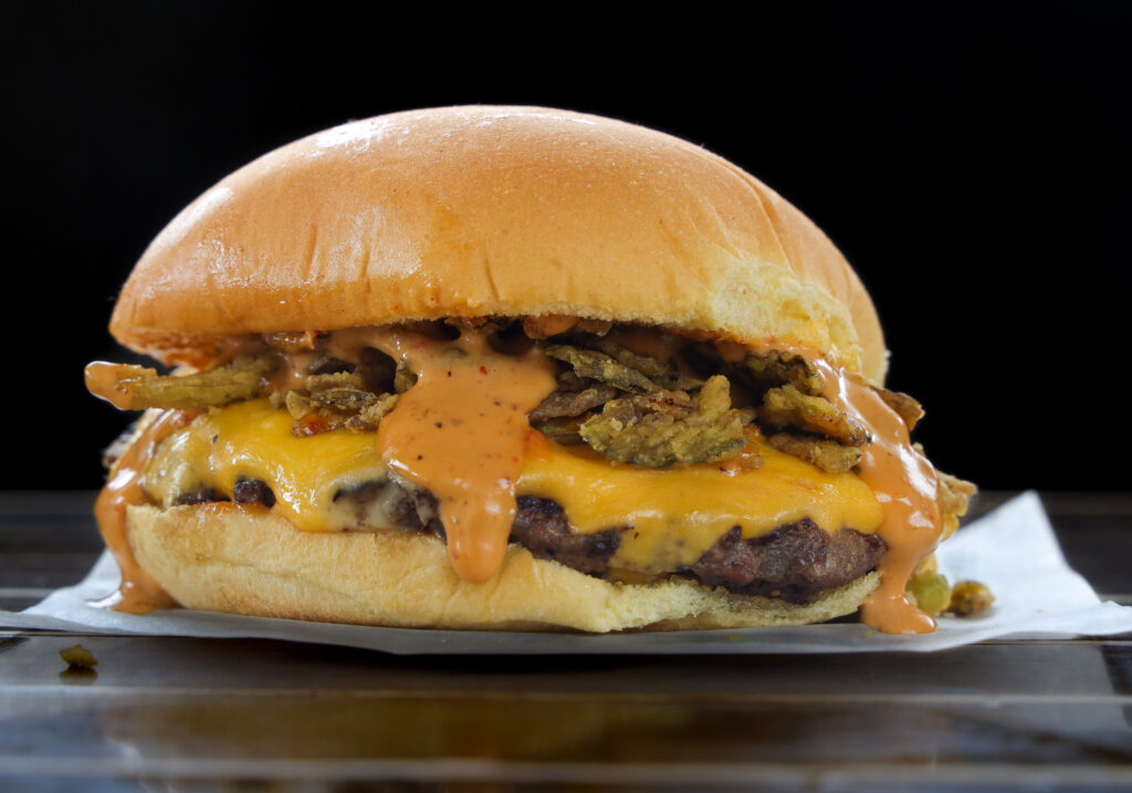 Double cheeseburger with melted american and pepper jack cheese, crispy fried jalapeños, and serrano hot sauce dripping onto a toasted potato bun