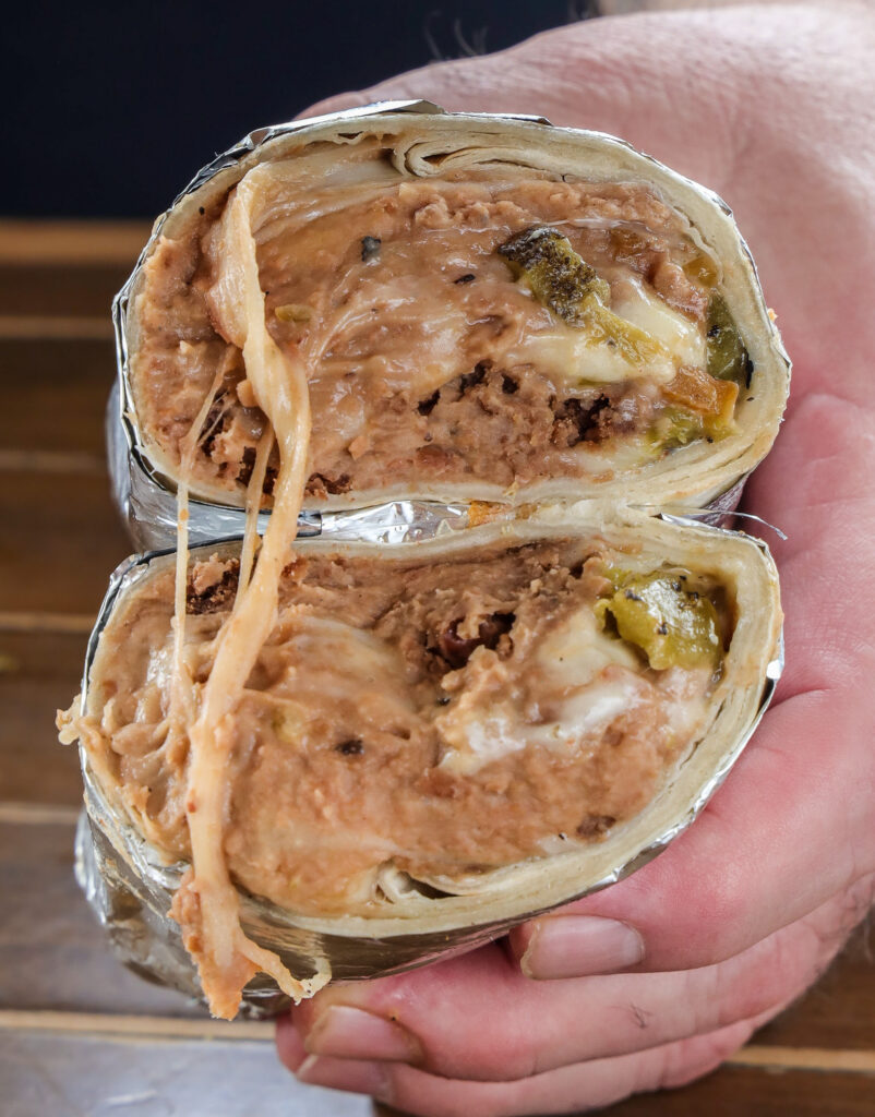 Bean and cheese burrito with roasted green chile sliced in half, showing melty cheese pull, held in hand